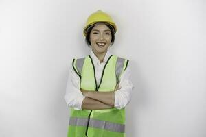 Smiling Asian woman labor worker in industry factory, posing with arms folded, wearing yellow safety helmet, green vest and uniform, isolated white background. photo