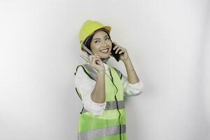 A smiling Asian woman labor wearing safety helmet and vest, having a phone call and pointing to copy space beside her, isolated by white background. Labor's day concept. photo