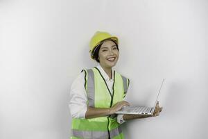 un sonriente asiático mujer labor vistiendo la seguridad casco y chaleco mientras participación su computadora portátil, aislado por blanco antecedentes. mano de obra día concepto. foto