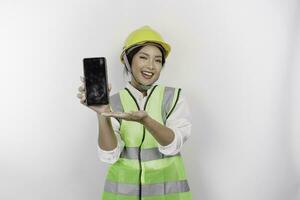 A smiling Asian woman labor wearing safety helmet and vest while showing her phone screen, isolated by white background. Labor's day concept. photo
