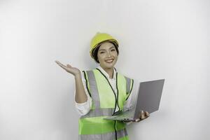 A smiling Asian woman labor wearing safety helmet and vest, pointing to copy space above her and holding her laptop, isolated by white background. Labor's day concept. photo