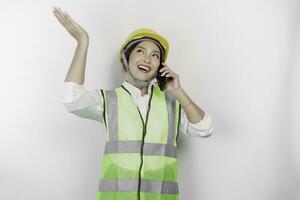 A smiling Asian woman labor wearing safety helmet and vest, talking on her phone and pointing to copy space above her, isolated by white background. Labor's day concept. photo