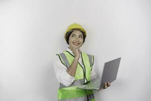 un pensativo joven mujer labor trabajador vistiendo la seguridad casco y chaleco mientras participación su ordenador portátil y mirando aparte a Copiar espacio, aislado por blanco antecedentes. mano de obra día concepto. foto