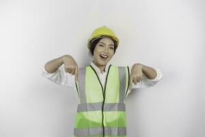 A smiling Asian woman labor wearing safety helmet and vest, pointing to copy space below her, isolated by white background. Labor's day concept. photo