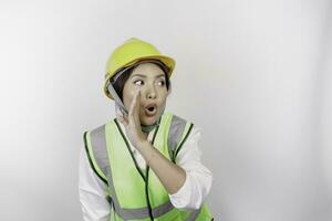 Young beautiful woman labor wearing safety helmet and vest is shouting and screaming loud with a hand on her mouth. Labor's day concept. photo