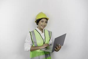 un sonriente asiático mujer labor vistiendo la seguridad casco y chaleco mientras participación su computadora portátil, aislado por blanco antecedentes. mano de obra día concepto. foto