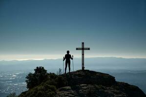 Woman contemplating a backlit landscape from the top of a hill, where there is a cross. photo