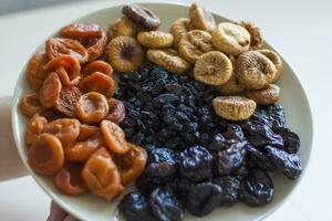 de los hombres manos sostener un plato de seco Fruta terminado el mesa. ciruelas pasas, seco albaricoques, higos, Pasas en un grande plato. foto