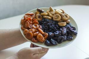 de los hombres manos sostener un plato de seco Fruta terminado el mesa. ciruelas pasas, seco albaricoques, higos, Pasas en un grande plato. foto