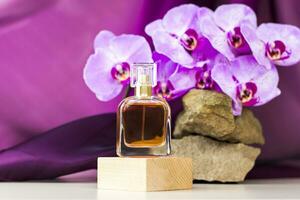 The perfume bottle stands on a wooden pedestal against a background of stone and beautiful orchids. Stylish appearance of the product, layout, personality. Banner, a place for the text. photo