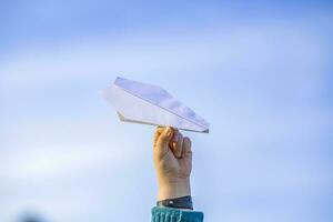 el Niños mano sostiene un blanco papel avión en contra el cielo. foto