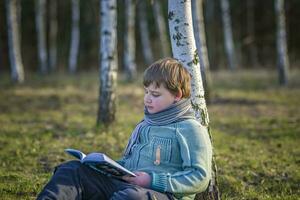 un hermoso chico en un bufanda es en un primavera parque, sentado debajo un abedul árbol, leyendo un libro y disfrutando su Sueños. foto