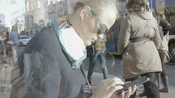 blond femme en utilisant mobile téléphone contre verre réflexion de gens en mouvement le long de le rue video