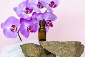 A brown bottle of cosmetic oil stands on a large structural stone with beautiful orchids next to it. Stylish appearance of the product, layout, personality. photo