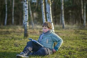 A handsome boy in a scarf is in a spring park, sitting under a birch tree, reading a book and enjoying his dreams. photo