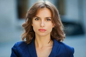 Portrait of a beautiful young woman with long brown hair, dressed in a blue jacket, standing against the background of an office building. Portrait of young business woman using mobile phone photo