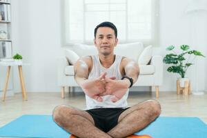 Online class training. Asian mature man stretching in sportswear making stretching for legs on mat yoga in living room interior and smiling. Happy sportsman workout by him slef. Sport at home. photo