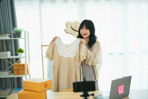 Asian woman steamming sell cloth shirt product online live at home. Young woman using mobile phone video call shows goods to customer and detail. Shopping online e-commerce buying and selling concept. photo