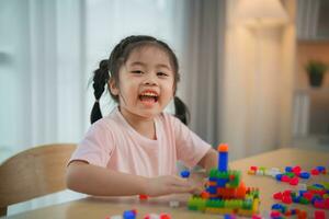 alegre asiático niña contento y sonriente jugando vistoso Lego juguetes, sentado en el mesa en el vivo habitación, creativamente jugando con Lego, edificio vistoso estructuras creatividad imaginar. foto