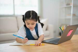 Asian baby girl smiling use laptop or writing drawing color on note book study online on wood table desk in living room at home. Education learning online from home concept. photo