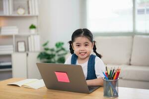 asiático bebé niña sonriente utilizar ordenador portátil o escritura dibujo color en Nota libro estudiar en línea en madera mesa escritorio en vivo habitación a hogar. educación aprendizaje en línea desde hogar concepto. foto