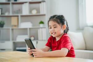 Asian child girl looking using and touch mobile phone screen. Baby smiling funny time to use mobile phone. Too much screen time. Cute girl watching videos while tv in living room, Internet addiction. photo