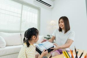 Grateful asian girl holding blue present box giving mothers day gift to mom. Happy mom and cute daughter kid child celebrating birthday, hugging on couch sofa at home. Happy family concept. photo