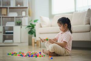 alegre asiático niña contento y sonriente jugando vistoso Lego juguetes, sentado en el vivo habitación piso, creativamente jugando con Lego, edificio vistoso estructuras creatividad imaginar. aprendizaje educación. foto