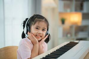 Asian cute girl smile and wearing white headphone playing learning online piano music in the living room at home. The idea of activities for the child at home during quarantine. Music learning study. photo