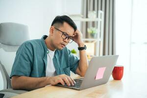 Asian Business man wear glasses and hand over head having stressful depression sad time while working on laptop at home. Depression man sad serios working from home. Working at anywhere concept. photo