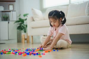 alegre asiático niña contento y sonriente jugando vistoso Lego juguetes, sentado en el vivo habitación piso, creativamente jugando con Lego, edificio vistoso estructuras creatividad imaginar. aprendizaje educación. foto