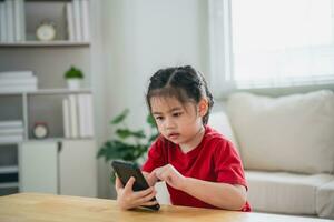 Asian child girl looking using and touch mobile phone screen. Baby smiling funny time to use mobile phone. Too much screen time. Cute girl watching videos while tv in living room, Internet addiction. photo