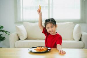 asiático contento niños niña comiendo Pizza y sonriente en el vivo habitación a hogar. niños niña comiendo y saboreo italiano hecho en casa Pizza en casa. foto