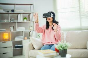 Asian woman smile and play VR game, 3D divice new innovation glasses for entertain in living room at home, asian woman joyful in house on holiday. Happy woman playing metaverse VR technology concept. photo