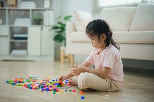 alegre asiático niña contento y sonriente jugando vistoso Lego juguetes, sentado en el vivo habitación piso, creativamente jugando con Lego, edificio vistoso estructuras creatividad imaginar. aprendizaje educación. foto