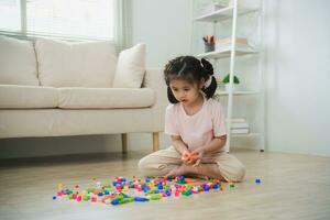 alegre asiático niña contento y sonriente jugando vistoso Lego juguetes, sentado en el vivo habitación piso, creativamente jugando con Lego, edificio vistoso estructuras creatividad imaginar. aprendizaje educación. foto