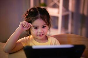 Asian girl smiling, laughing, excited, looking, wearing white headphone and using  pen touch on white screen tablet screen streaming night light. Cute girl watching videos while tv Internet addiction. photo