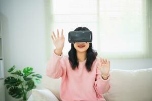 Asian woman smile and play VR game, 3D divice new innovation glasses for entertain in living room at home, asian woman joyful in house on holiday. Happy woman playing metaverse VR technology concept. photo