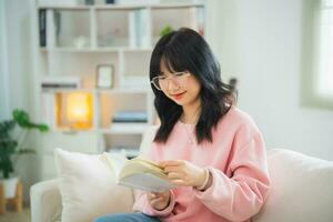 alegre contento asiático mujer sonriente y leyendo libro mientras sentado en sofá sofá en vivo habitación a hogar. asiático mujer relajarse leyendo libro sonrisa a sofá en el casa. actividad pasatiempo a casa concepto. foto