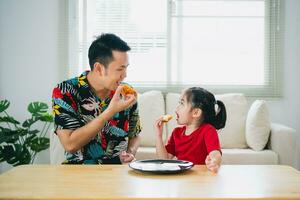 asiático contento niños niña y su padre alimentación comiendo Pizza y sonriente en el vivo habitación a hogar. niños niña y su papá comiendo y saboreo italiano hecho en casa Pizza en casa. foto