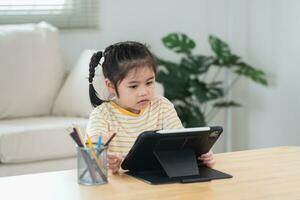 Asian girl looking and touch on white screen at the tablet screen attentively. overstimulated children concept. Too much screen time. Cute girl watching videos while tv, Internet addiction concept. photo