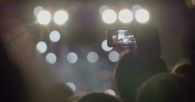 Spectator man recording video of stage in front of bright spotlights via smartphone at outdoor music concert