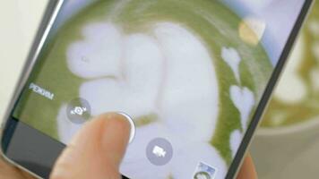 On the table is a cup of coffee with foam and pattern and girl takes photo of cup on mobile phone video