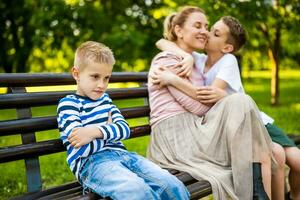 Happy mother is sitting with her sons on bench in park. One boy is offended and pouting. photo