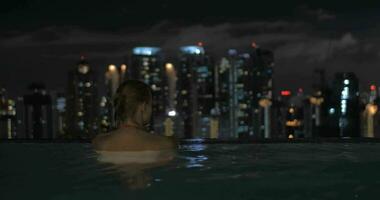 View of woman in the pool on the skyscraper roof and then swimming man about her on night city landscape Kuala Lumpur, Malaysia video