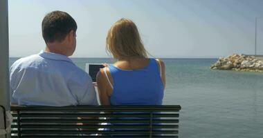 Jeune femme et homme sont séance sur banc sur plage sur mer horizon Contexte en train de regarder quelque chose dans tablette ordinateur et Parlant le Pirée, Grèce video