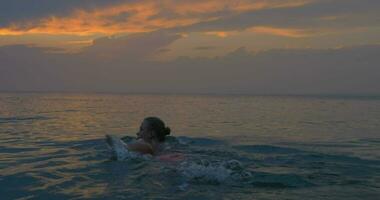 joven mujer nada lejos en el mar a atardecer, mágico ver de noche nublado cielo y calma mar video