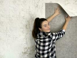Happy brunette girl making interior change. Applying new gray wallpaper on white wall in room. Repair work of home. photo