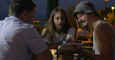 Group of friends sitting in the evening at the table and watches videos on the tablet, one of the men, talking and gesticulating