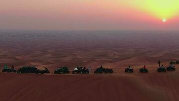 un zumbido moscas terminado vehículos todo terreno en pie en el arena dunas de el Desierto en contra el fondo de el puesta de sol. aéreo ver video